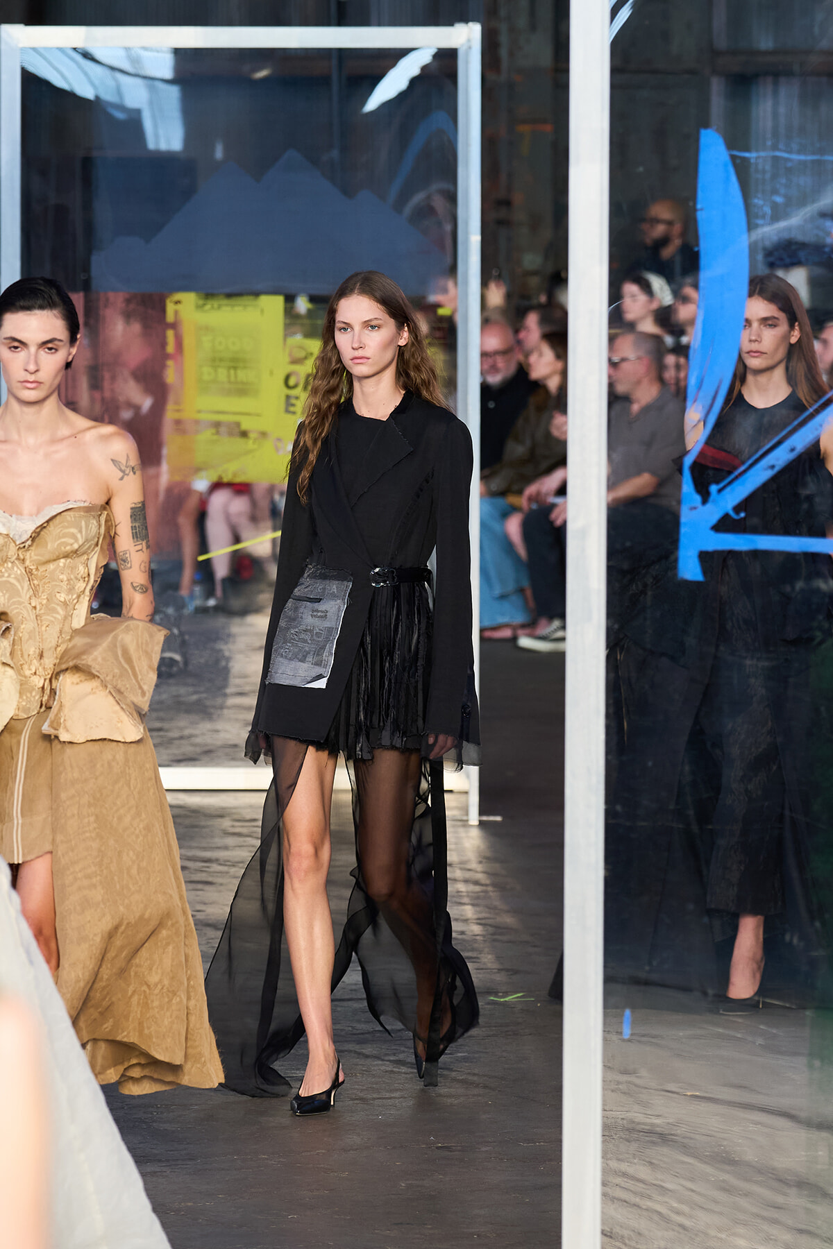 Model walks a runway wearing a black blazer and sheer black skirt as onlookers sit behind glass panels in the background, left model in a beige gown nearby.