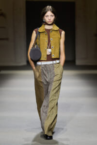 Model walking runway in mustard suede vest over brown top with wide plaid trousers and a round gray bag slung over the shoulder
