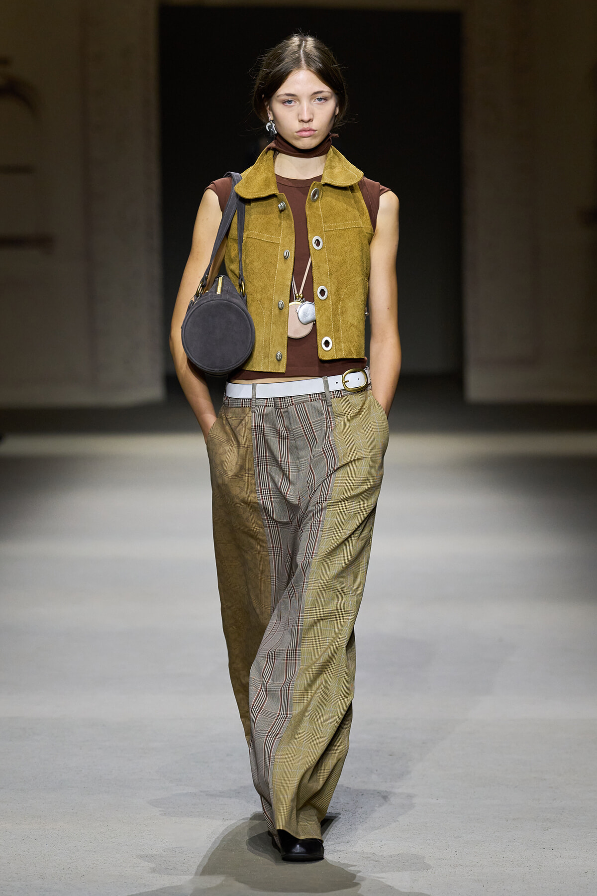 Model walking runway in mustard suede vest over brown top with wide plaid trousers and a round gray bag slung over the shoulder
