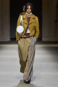 Model on the runway wearing a mustard suede jacket over a brown top, with oversized plaid pants and a round white handbag.