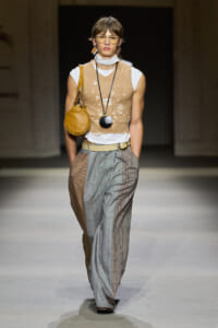 Model walking the runway in a beige distressed knit vest over a white top, paired with patchwork gray and brown plaid wide-leg trousers and a tan shoulder bag.