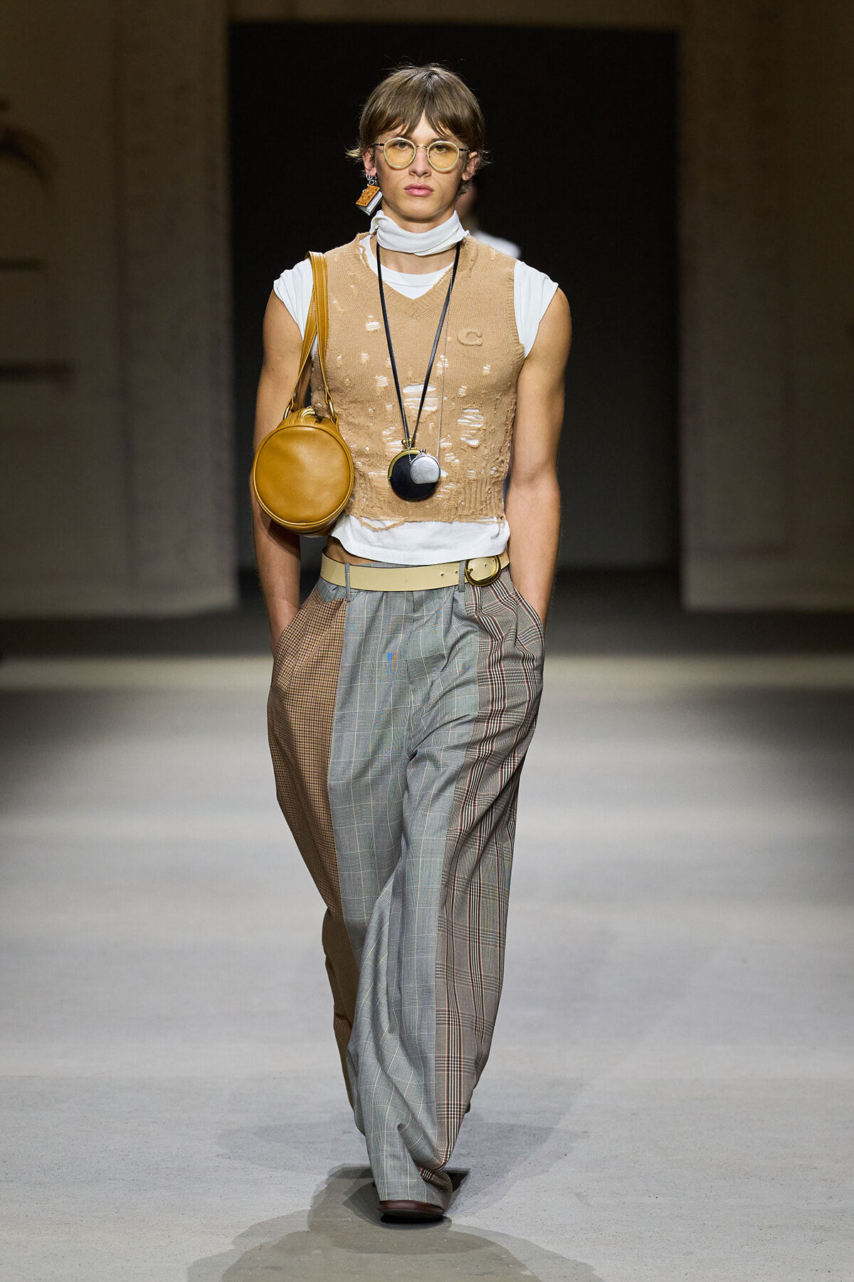 Model walking the runway in a beige distressed knit vest over a white top, paired with patchwork gray and brown plaid wide-leg trousers and a tan shoulder bag.