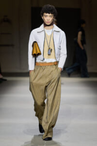 Male model on a runway wearing tan plaid wide trousers, a white jacket, beige layered top, brown belt, and layered necklaces with a brown clutch under the left arm.