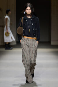Model walks the runway in a dark cropped jacket, black top, and tan plaid wide-leg trousers with a brown belt and round brown bag over the shoulder