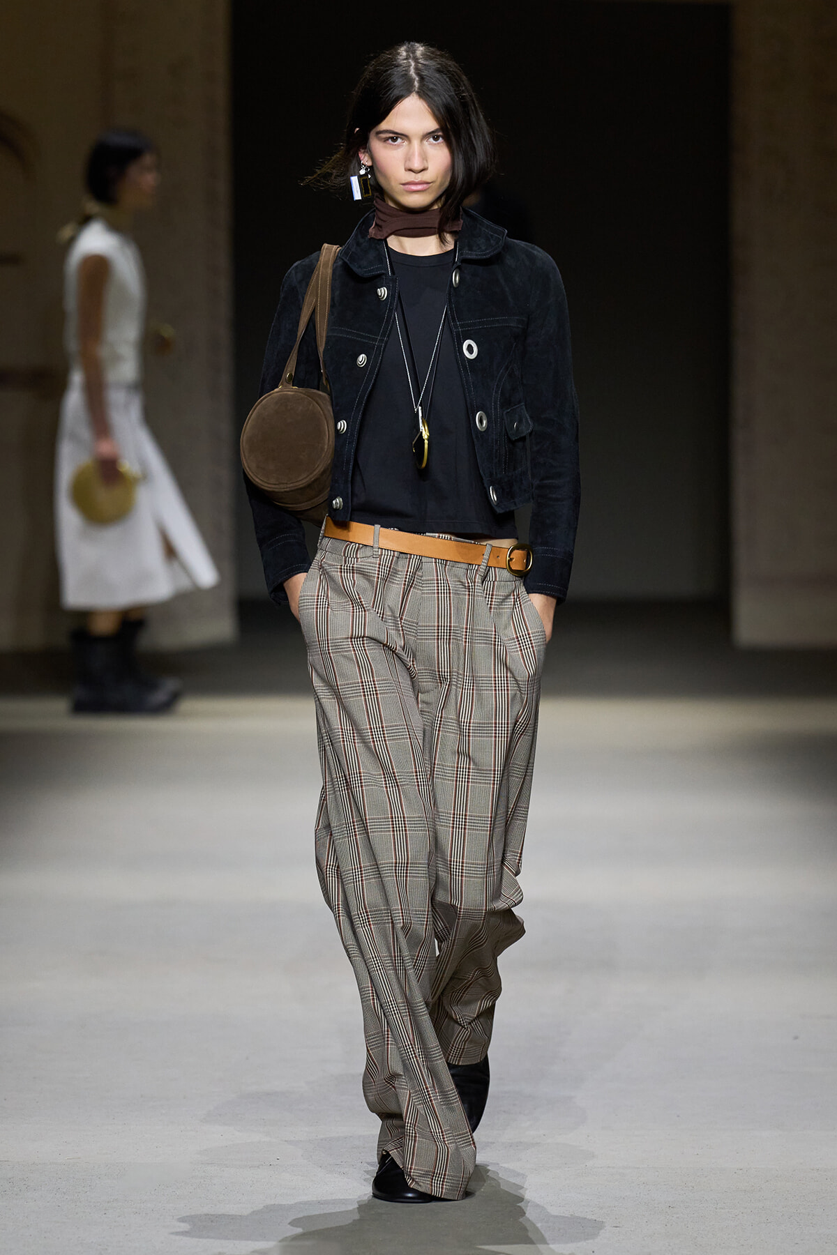 Model walks the runway in a dark cropped jacket, black top, and tan plaid wide-leg trousers with a brown belt and round brown bag over the shoulder