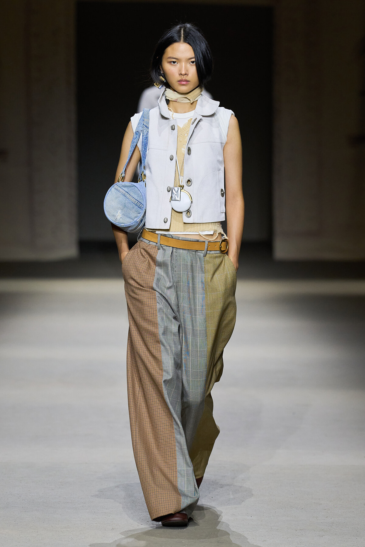 Model on a runway wearing color-block patchwork wide-leg pants, a light sleeveless vest, beige scarf, and a round blue handbag.