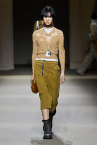 Model walks a runway in a beige distressed knit sweater over a white top, olive midi skirt with zipper, black boots, and a brown bag.