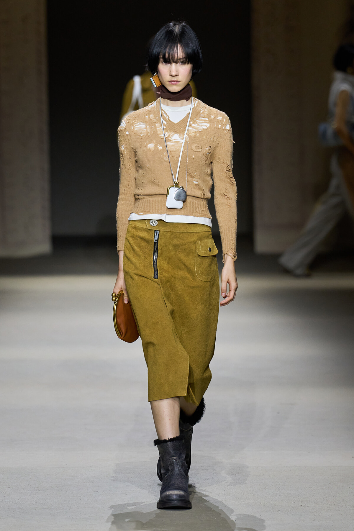 Model walks a runway in a beige distressed knit sweater over a white top, olive midi skirt with zipper, black boots, and a brown bag.