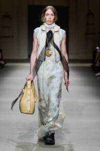 Model walking a runway in a pale blue, cream dress with cloud appliqués, a cream vest, sheer scarf, and a large yellow handbag