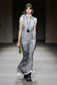 Model walking runway in a floor-length silver-gray cityscape print dress, white scarf, long black pendant necklace, and black boots, carrying a small round tan bag