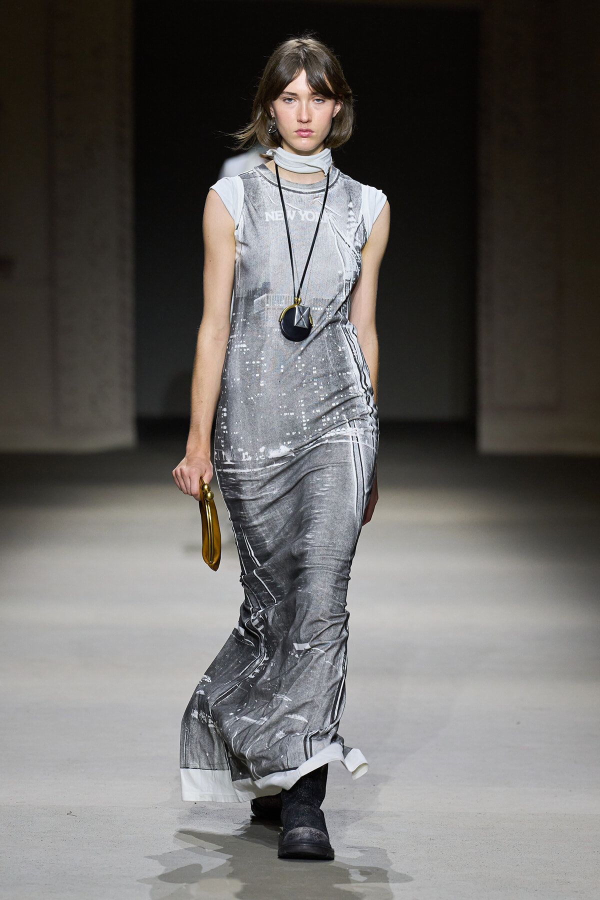 Model walking runway in a floor-length silver-gray cityscape print dress, white scarf, long black pendant necklace, and black boots, carrying a small round tan bag