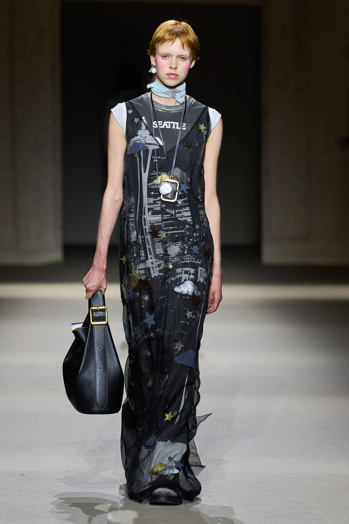 Model walking a fashion runway in a sheer black star-printed gown with the word 'SEATTLE', layered necklaces, and a light blue scarf at the neck.