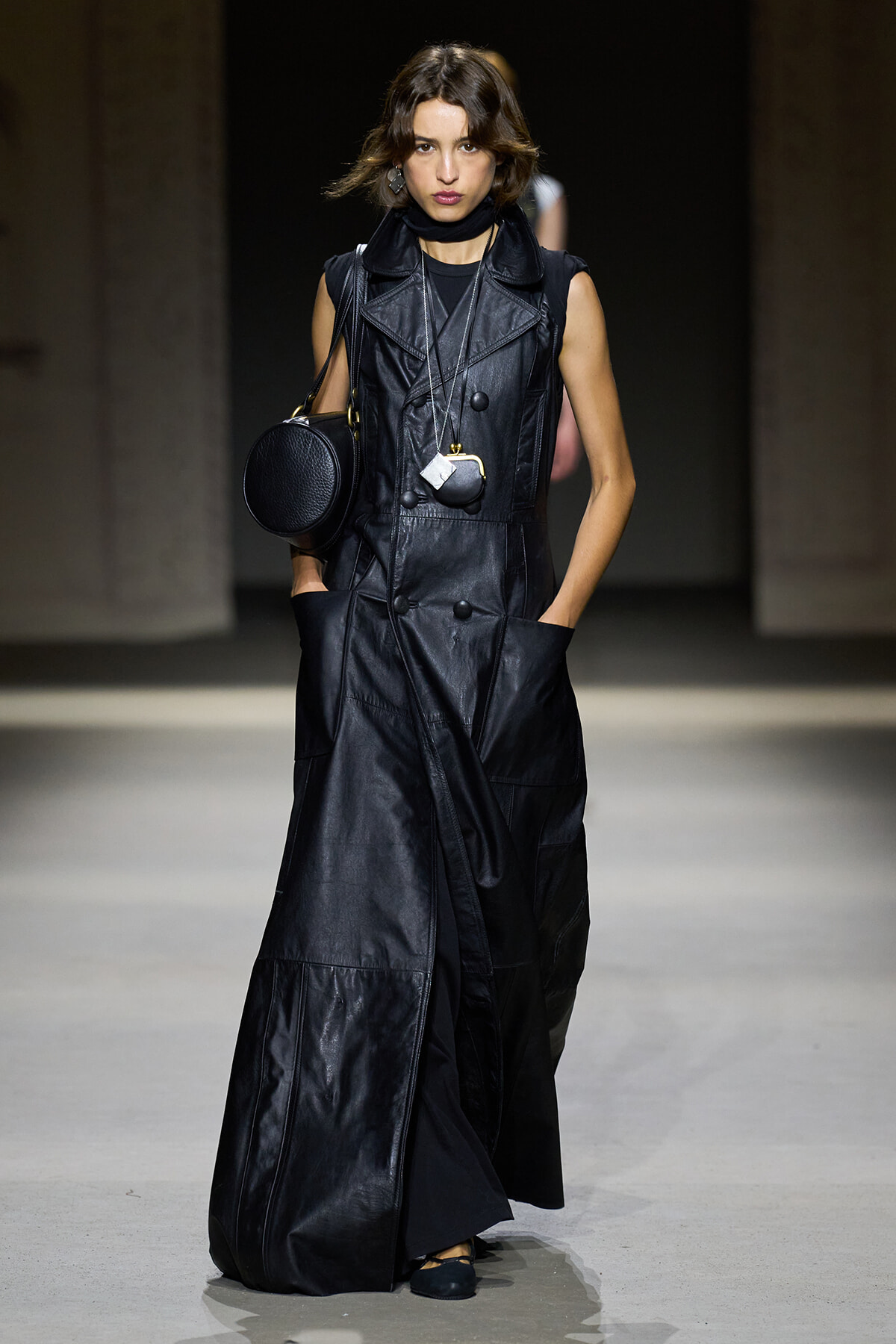 Model walking a runway in a long black leather sleeveless trench with large pockets, paired with a circular black bag and layered necklaces.