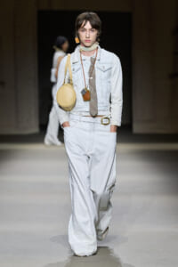 Model walking runway in a light denim jacket and wide-leg white pants, accessorized with a circular beige bag and layered necklaces.