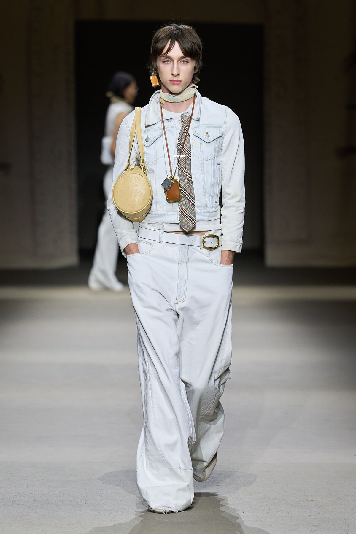 Model walking runway in a light denim jacket and wide-leg white pants, accessorized with a circular beige bag and layered necklaces.