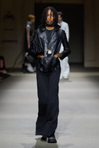 Model walking a runway in a black leather blazer, black wide-leg pants, and a long pendant necklace with a small pouch.