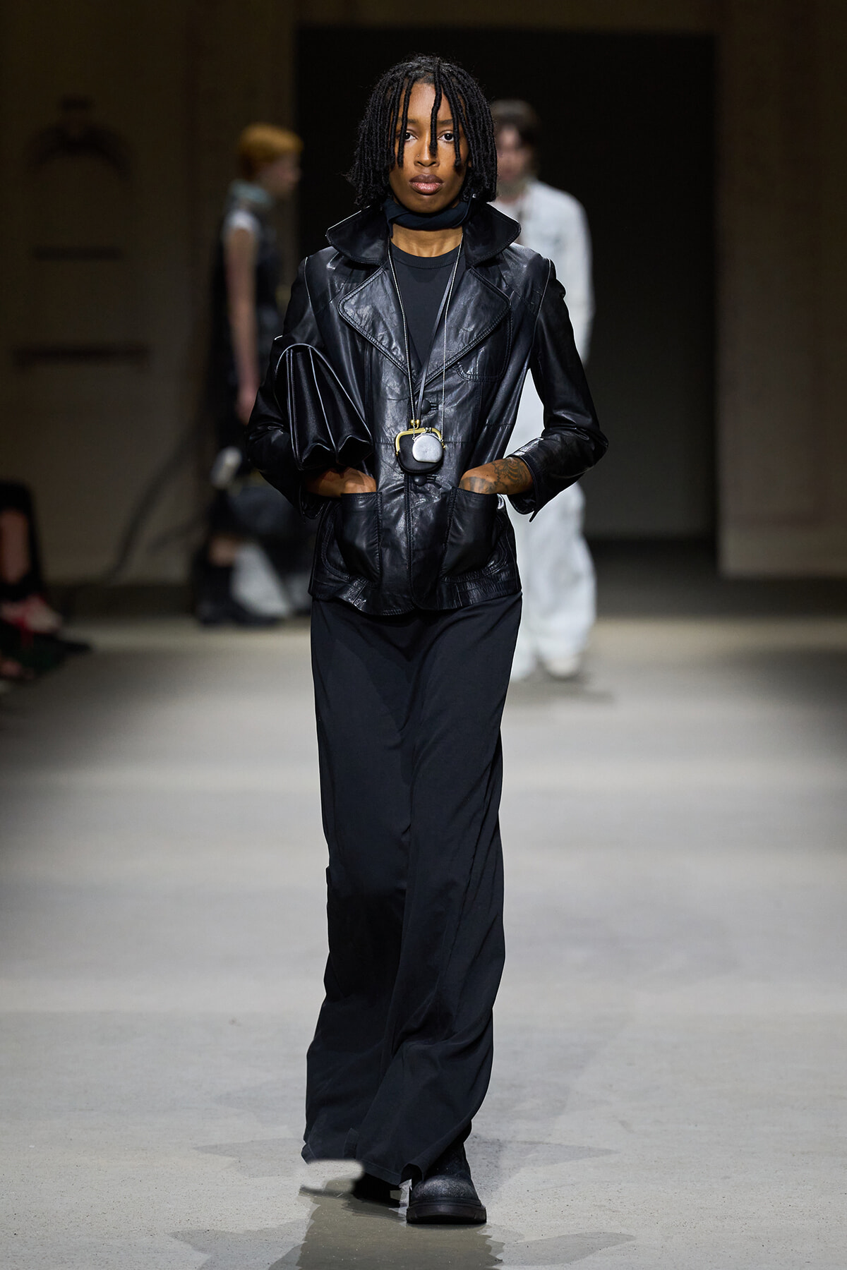 Model walking a runway in a black leather blazer, black wide-leg pants, and a long pendant necklace with a small pouch.