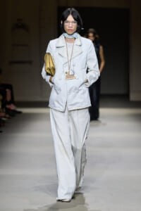 Model walks the runway in an oversized white suit with wide-leg pants, white-framed glasses, and a yellow clutch accessory.