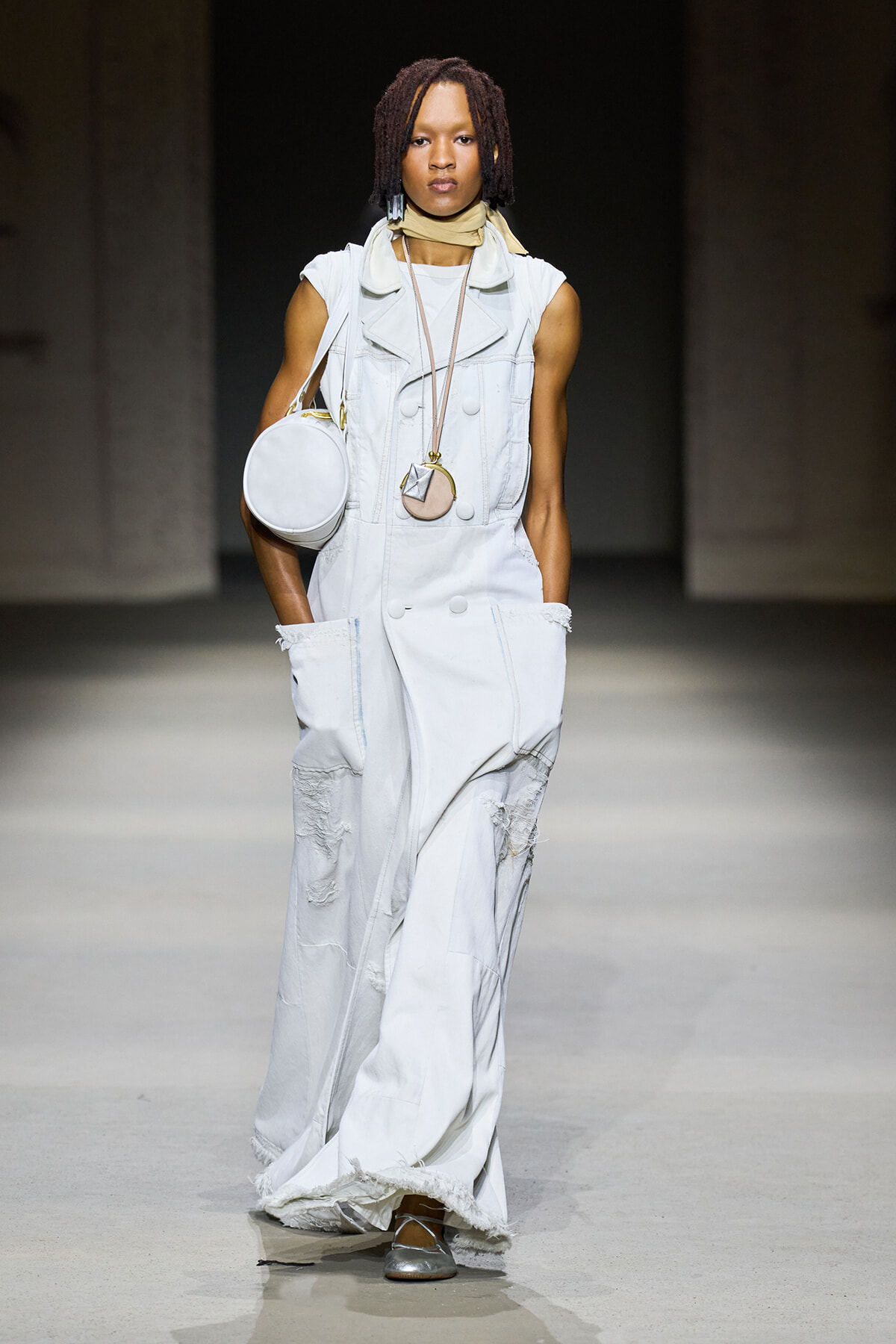Model walking the runway in a sleeveless white distressed denim jumpsuit with large pockets, paired with a round white bag and layered necklaces, on a minimalist catwalk.