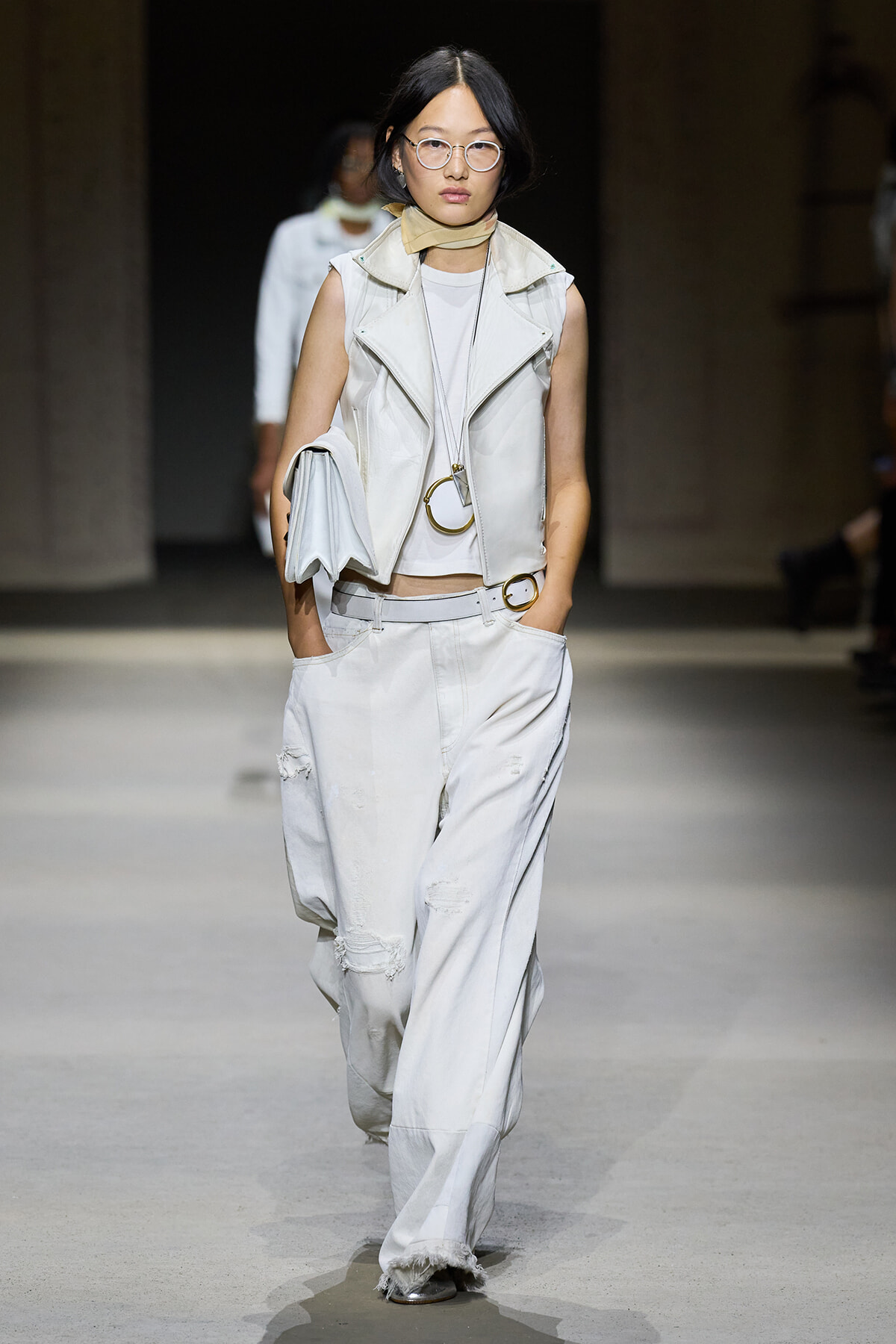 Female fashion model on a runway wearing an all-white ensemble: sleeveless leather vest over a white top, wide-leg distressed white jeans, beige scarf, round glasses, and a white clutch under one arm.