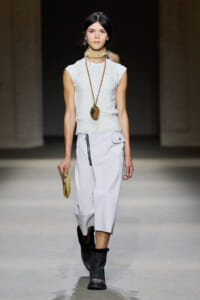Model on a runway wearing a white sleeveless knit top and midi skirt, beige neck scarf, long pendant necklace, black boots, and a small gold clutch.