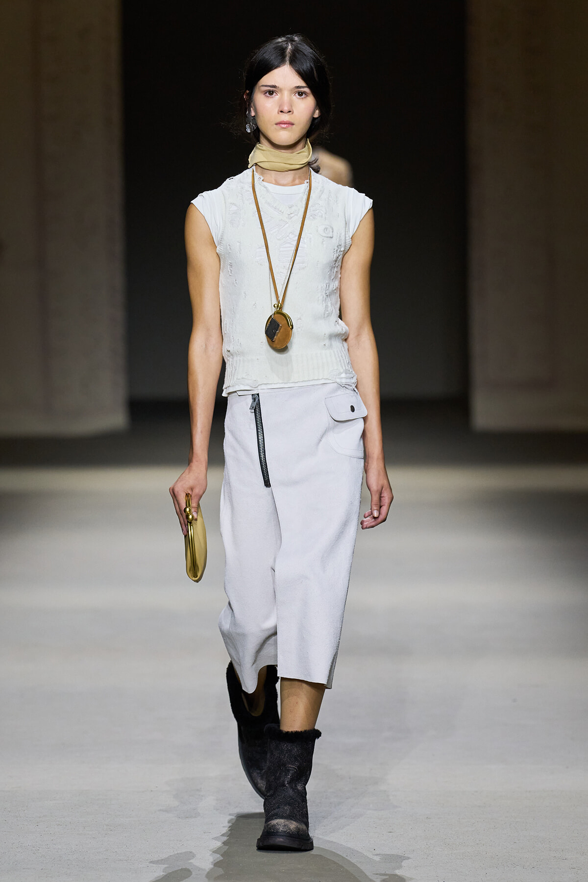Model on a runway wearing a white sleeveless knit top and midi skirt, beige neck scarf, long pendant necklace, black boots, and a small gold clutch.
