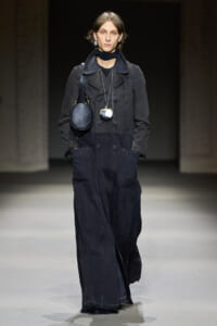 Model on a runway wearing a dark, oversized denim jumpsuit with a black scarf and two circular bags slung at the chest area.