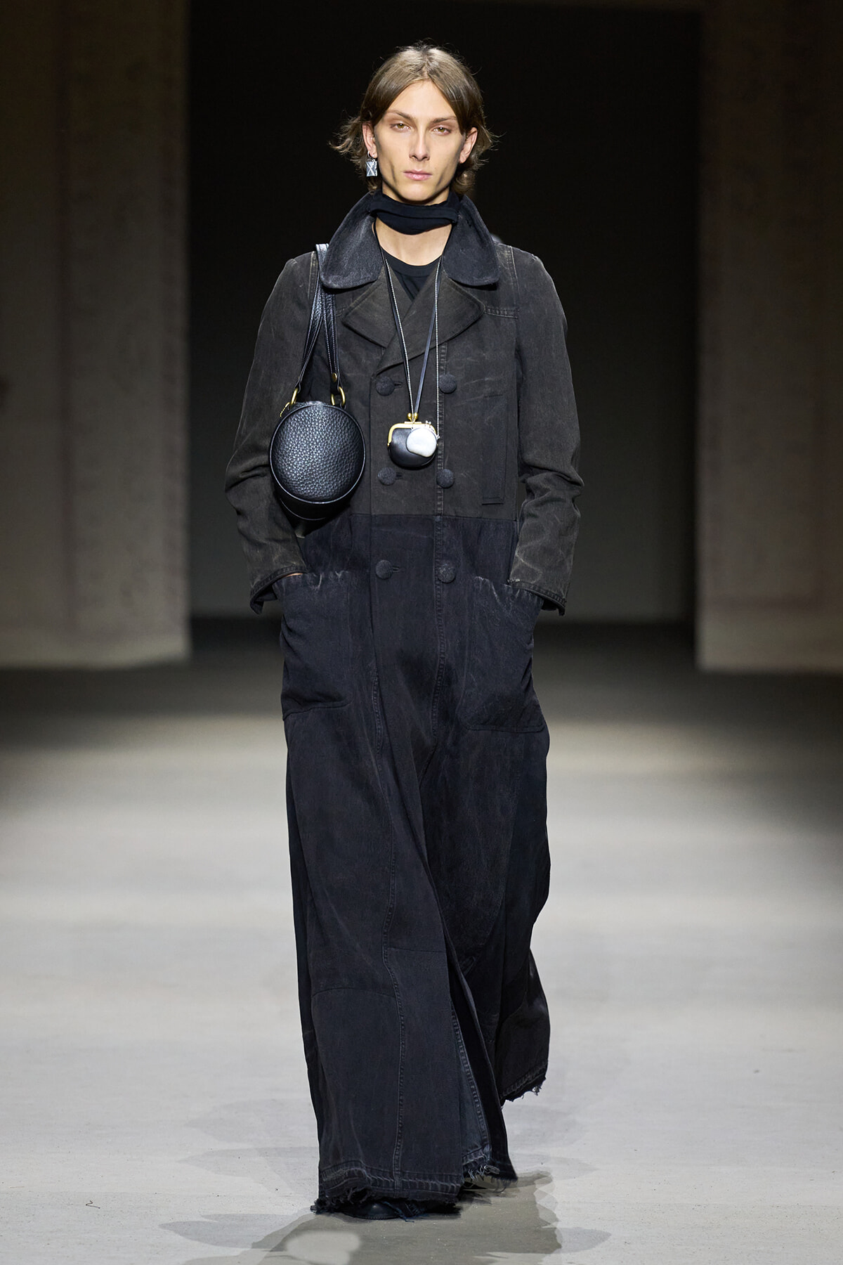 Model on a runway wearing a dark, oversized denim jumpsuit with a black scarf and two circular bags slung at the chest area.