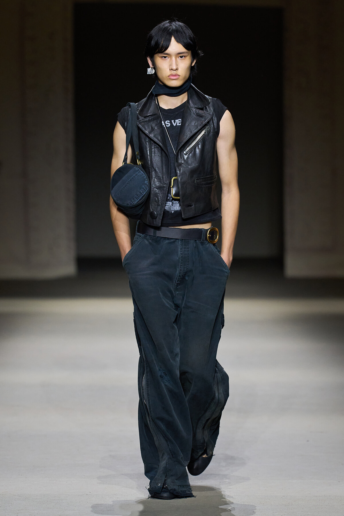 Male model on a runway wearing a black sleeveless leather vest, dark bag, and loose dark cargo pants with a round-buckle belt.