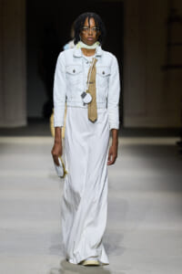 Model walking a runway in a white maxi dress with a light denim jacket, beige plaid necktie, scarf, and round glasses; holding a small circular purse and gold accessory.