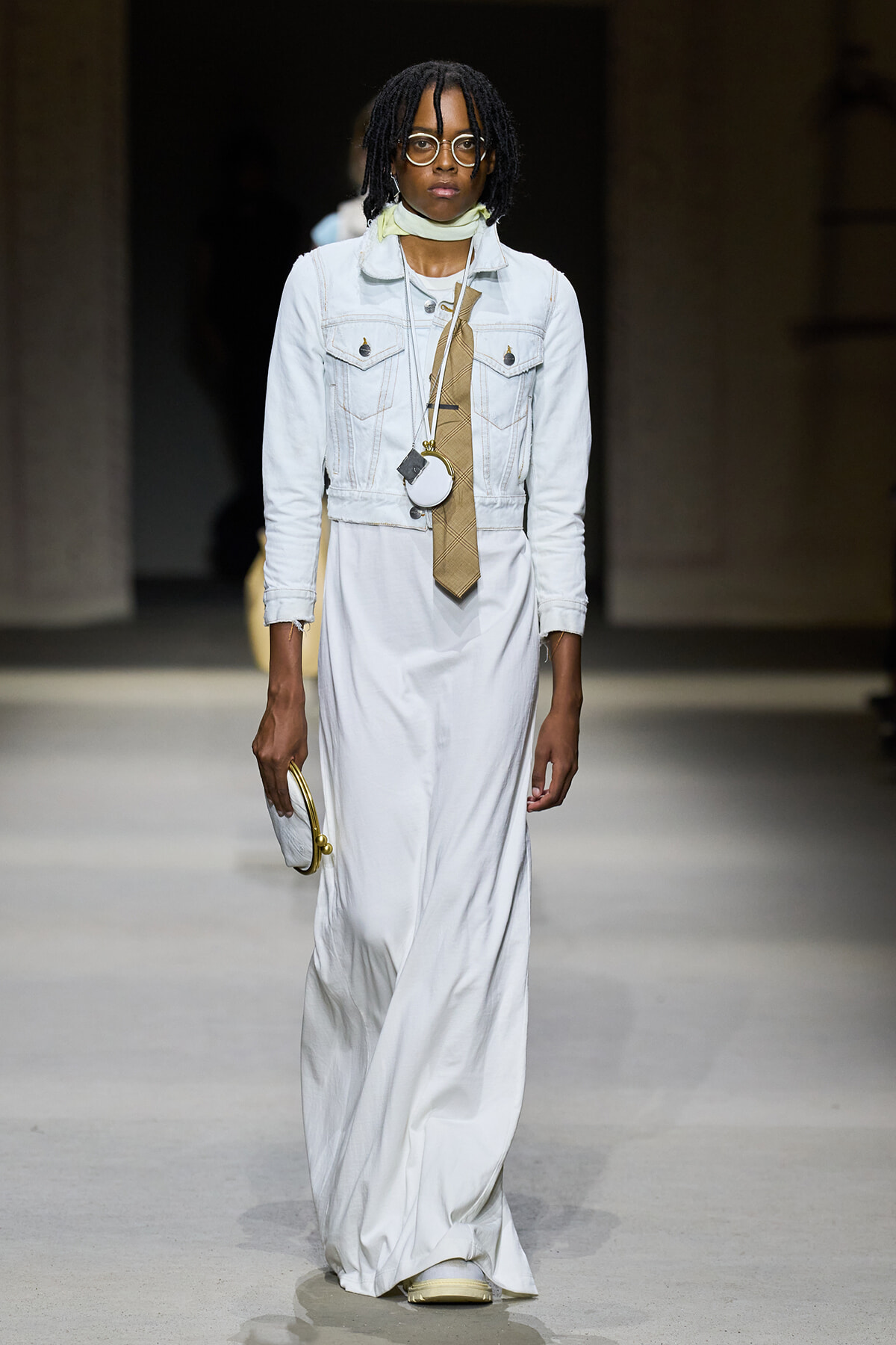 Model walking a runway in a white maxi dress with a light denim jacket, beige plaid necktie, scarf, and round glasses; holding a small circular purse and gold accessory.
