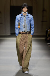 Runway model wearing a light denim jacket, gray tie, scarf, brown belt, and oversized brown patterned trousers with white sneakers and a small shoulder bag.