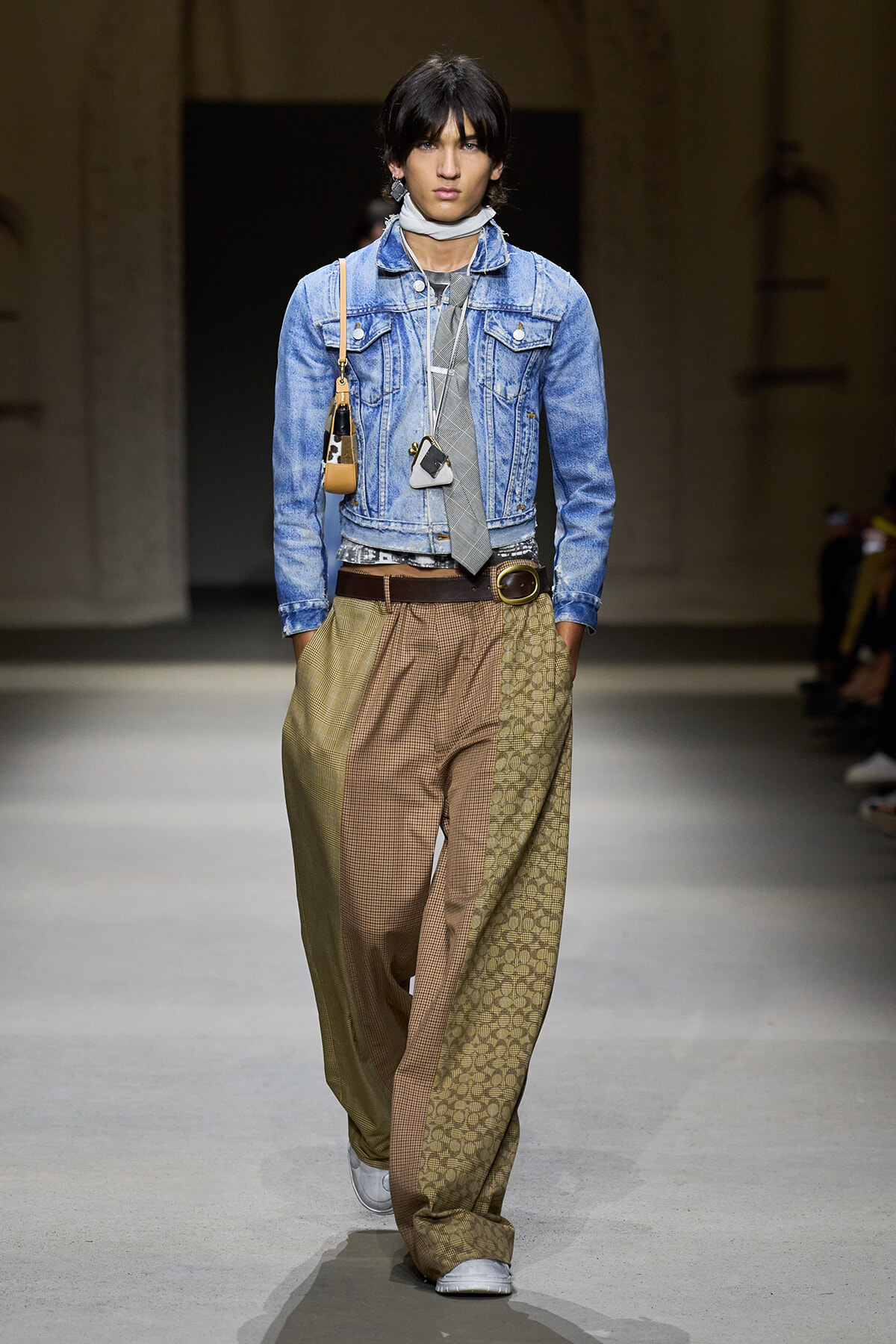 Runway model wearing a light denim jacket, gray tie, scarf, brown belt, and oversized brown patterned trousers with white sneakers and a small shoulder bag.