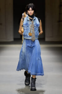 Runway model in a denim-on-denim outfit: sleeveless blue vest, long patchwork denim skirt, black top, plaid tie, black boots, and a small tan bag.