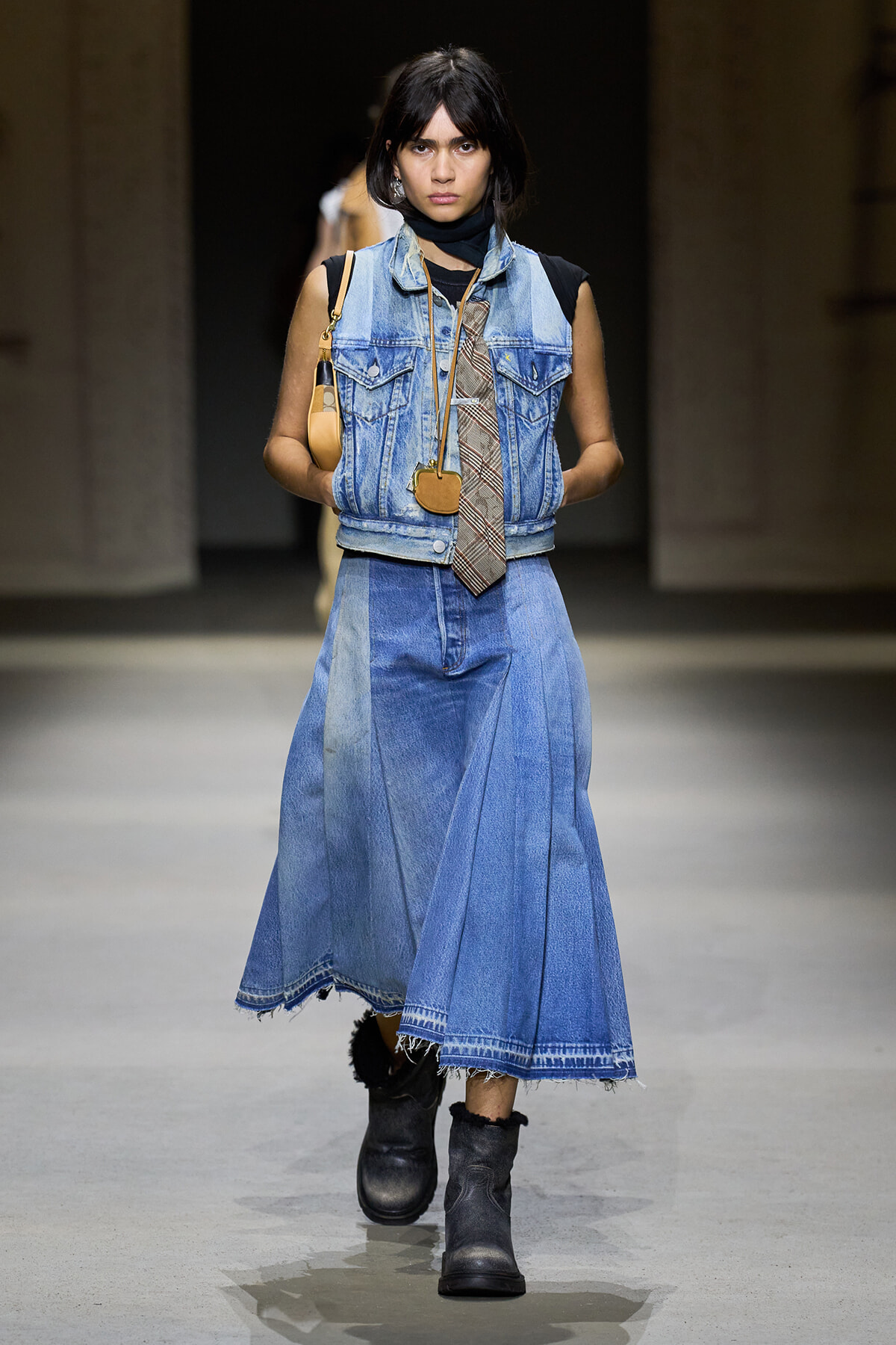 Runway model in a denim-on-denim outfit: sleeveless blue vest, long patchwork denim skirt, black top, plaid tie, black boots, and a small tan bag.