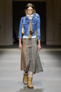 Runway model wearing a blue denim jacket over a white top and a long multi-pattern plaid skirt, tan boots, and yellow sunglasses.