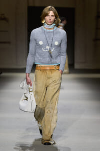 Model walking a runway in a light gray knit sweater, pale blue scarf, wide tan distressed pants with a tan belt, and a white handbag.