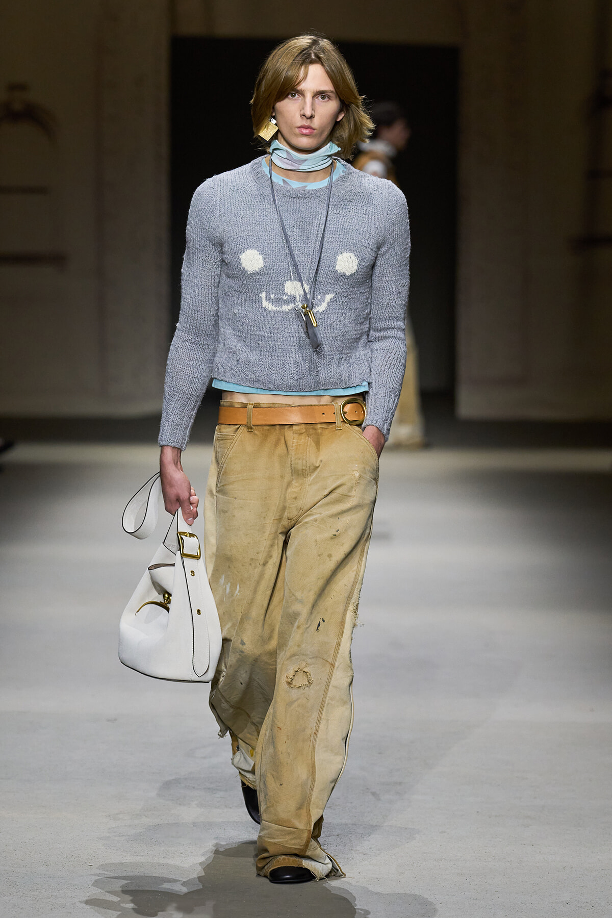 Model walking a runway in a light gray knit sweater, pale blue scarf, wide tan distressed pants with a tan belt, and a white handbag.