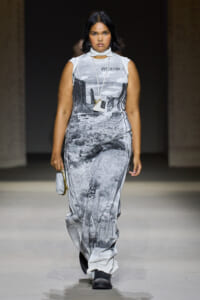 Model walking the runway in a sleeveless grayscale desert-print dress with a white neck scarf and a small white pendant bag.