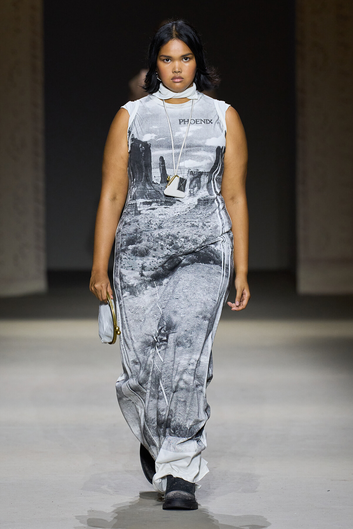 Model walking the runway in a sleeveless grayscale desert-print dress with a white neck scarf and a small white pendant bag.