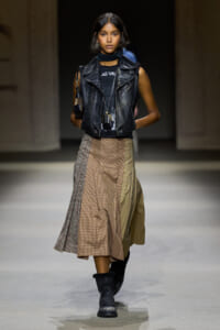 Model walks a fashion runway in a black leather biker vest over a dark top, with a scarf and a long plaid skirt paired with black boots.