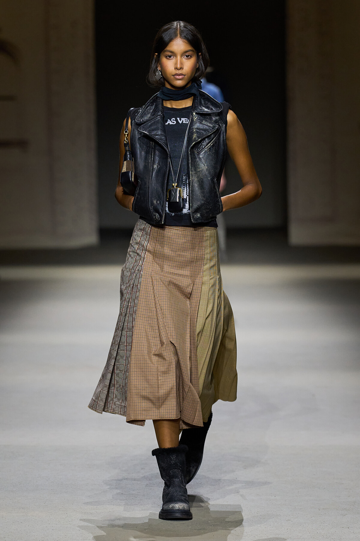 Model walks a fashion runway in a black leather biker vest over a dark top, with a scarf and a long plaid skirt paired with black boots.