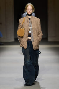 Model walking the runway in an oversized brown plaid blazer, striped tie, black turtleneck, and bag with distressed dark jeans and chunky accessories.