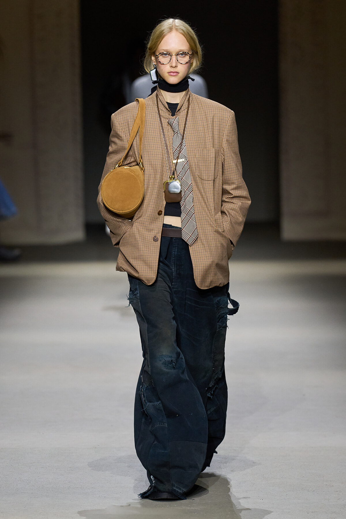 Model walking the runway in an oversized brown plaid blazer, striped tie, black turtleneck, and bag with distressed dark jeans and chunky accessories.