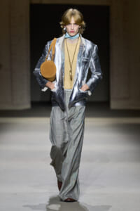 Model walking runway in metallic silver blazer, beige top, gray plaid wide-leg pants, with a round tan bag over the shoulder.