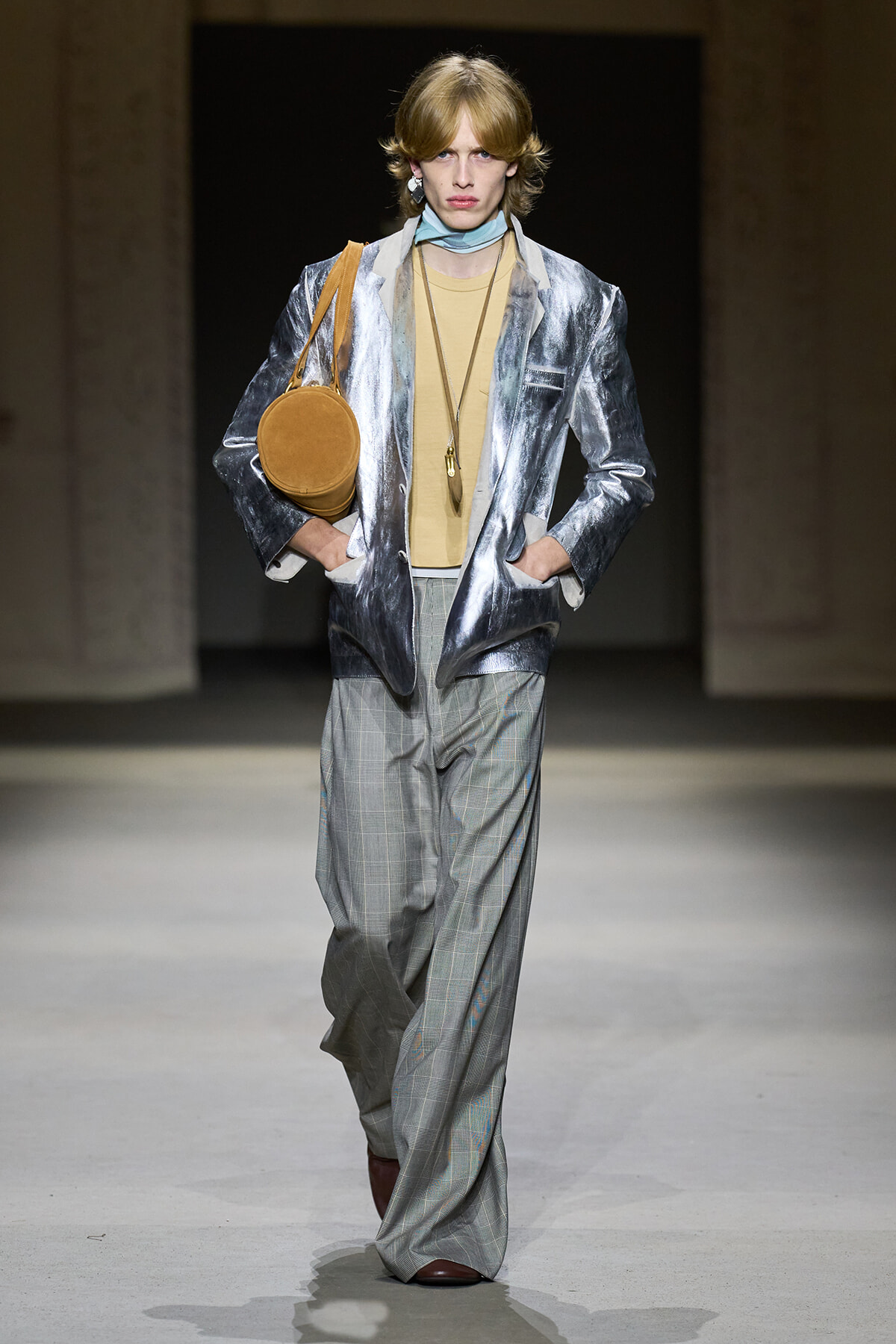 Model walking runway in metallic silver blazer, beige top, gray plaid wide-leg pants, with a round tan bag over the shoulder.