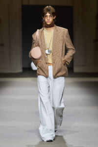Model on a runway wearing a beige check blazer over a tan top with white wide-leg pants, round glasses, and a pink circular bag.