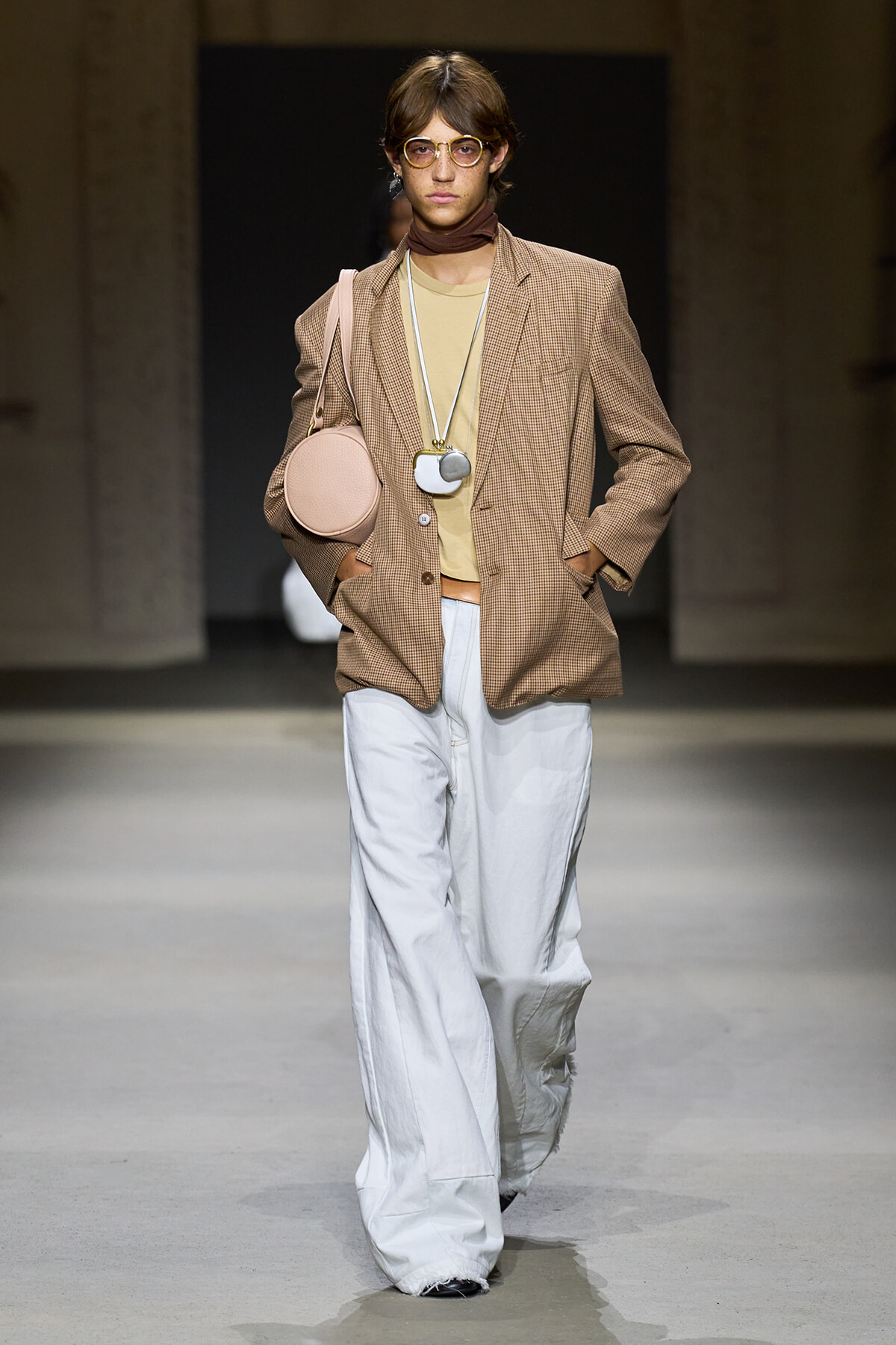 Model on a runway wearing a beige check blazer over a tan top with white wide-leg pants, round glasses, and a pink circular bag.
