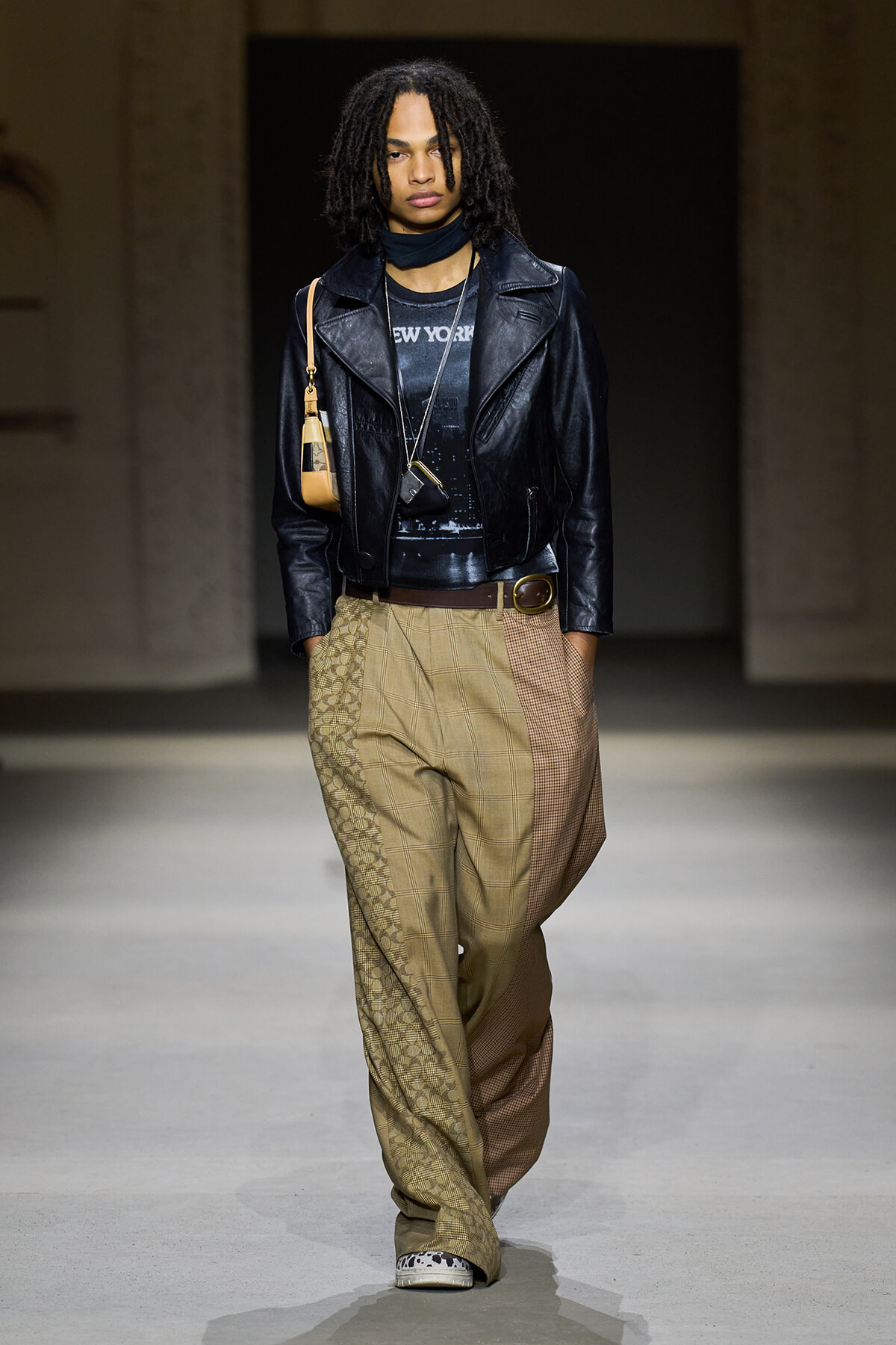 Model walking the runway in a black leather jacket over a dark graphic tee ('New York'), with oversized tan patchwork pants, brown belt, and a small beige shoulder bag.