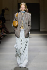 Model on a runway wearing a plaid blazer with pale blue wide-leg pants and a round tan shoulder bag, accessorized with a light scarf.